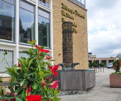 Une fontaine avec une sculpture devant un bâtiment moderne. Des fleurs violettes et rouges embellissent le premier plan.