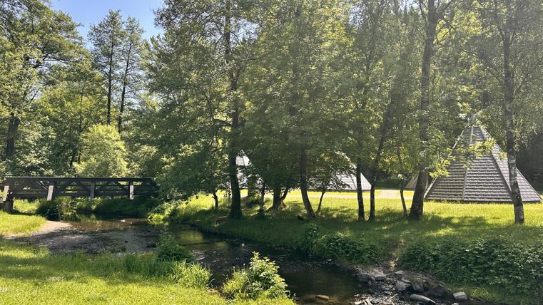 Een rustige landschap met een kleine rivier en brug. Op de achtergrond zijn bomen en piramidevormige gebouwen te zien.