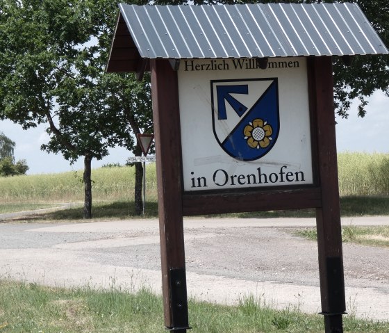 Ortsschild von Orenhofen mit Wappen, umgeben von B&auml;umen und Feldern. Ein Weg f&uuml;hrt durch die l&auml;ndliche Landschaft., &copy; TI Bitburger Land