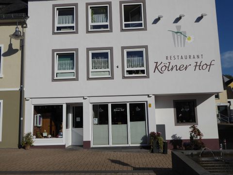 A restaurant named "Kölner Hof" with a bright facade and large windows. The surrounding area is paved, and there are some plants in front of the entrance.