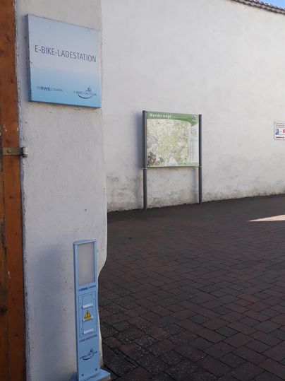 Een witte muur met een informatiebord en een kaartinformatie. De vloer is geplaveid en de lucht is blauw.