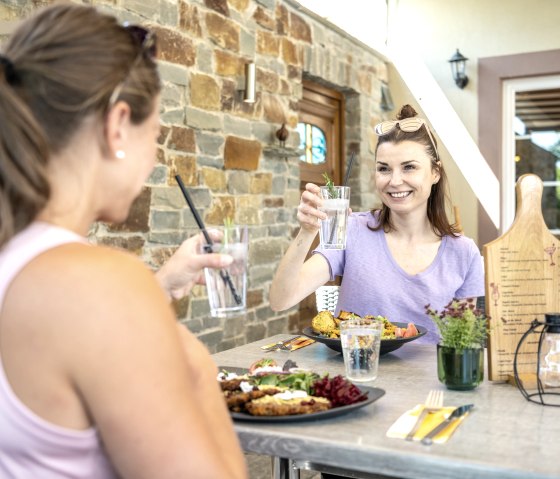 Twee vrouwen zitten aan een tafel buiten, proosten met een drankje en genieten van een maaltijd. Een stenen muur op de achtergrond., © Eifel Tourismus GmbH, Dominik Ketz