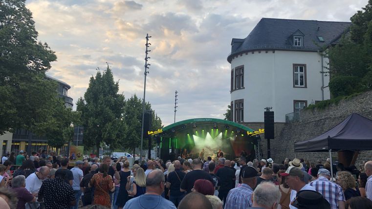 A crowd at an open-air concert in front of a stage. In the background, trees and buildings are visible, while the sky is partly cloudy.
