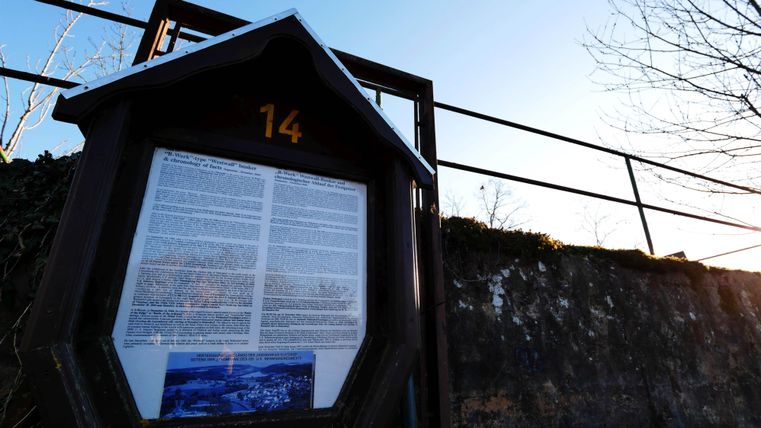 Un panneau d'information se trouve sur une pente. En arrière-plan, un ciel clair et quelques branches nues sont visibles.