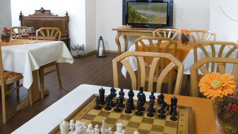 Ein gemütlicher Raum mit einem Schachbrett und einer dekorativen Blume auf dem Tisch. Im Hintergrund sind Tische und ein Fernseher sichtbar.