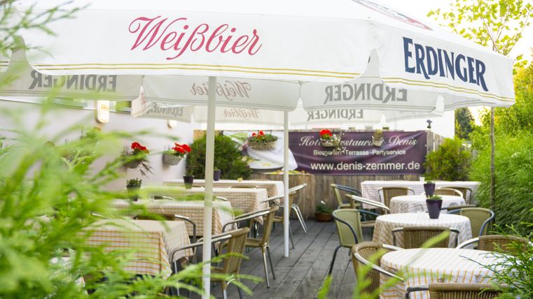 Een gezellige buitenruimte van een restaurant met tafels en stoelen. Een parasol met de opdruk "Weißbier" en "Erdinger" zorgt voor schaduw.
