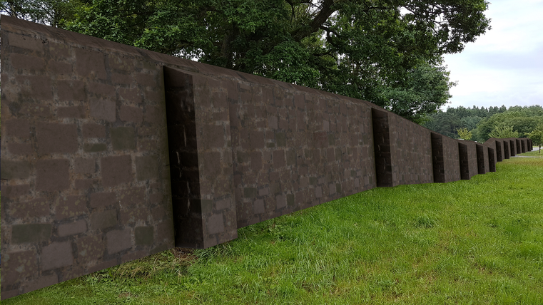 A long stone wall stands on a green meadow. In the background, trees and a clear sky are visible.