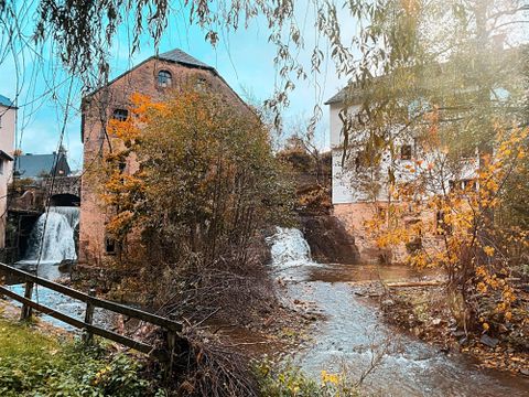 Eine malerische Landschaft mit alten Gebäuden und Wasserfällen. Die Bäume sind bunt gefärbt und das Wasser fließt ruhig vorbei.
