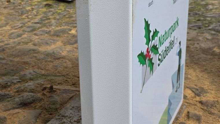 Een informatiedbord over de natuur in een parkachtige omgeving. Het bord heeft een heldere, witte oppervlakte met groene en kleurrijke symbolen.