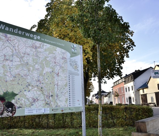 Carte de randonn&eacute;e dans le parc naturel S&uuml;deifel devant des arbres et des maisons automnales &agrave; Wolsfeld. On peut &eacute;galement y voir un banc de repos et une bo&icirc;te aux lettres., &copy; TI Bitburger Land