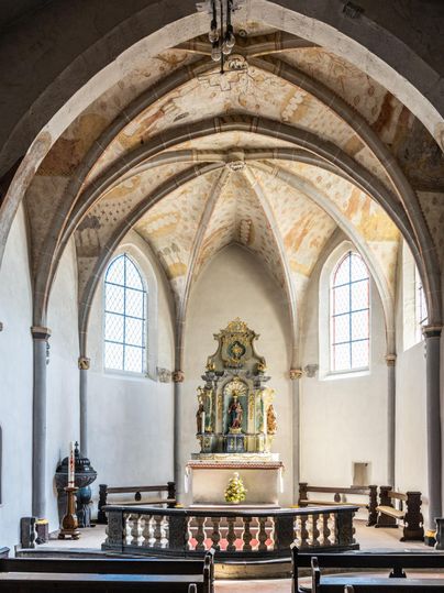 Der Innenraum einer Kirche mit hohen, gewölbten Decken und bunten Fenstern. Im Altarbereich befindet sich eine kunstvolle Statue.