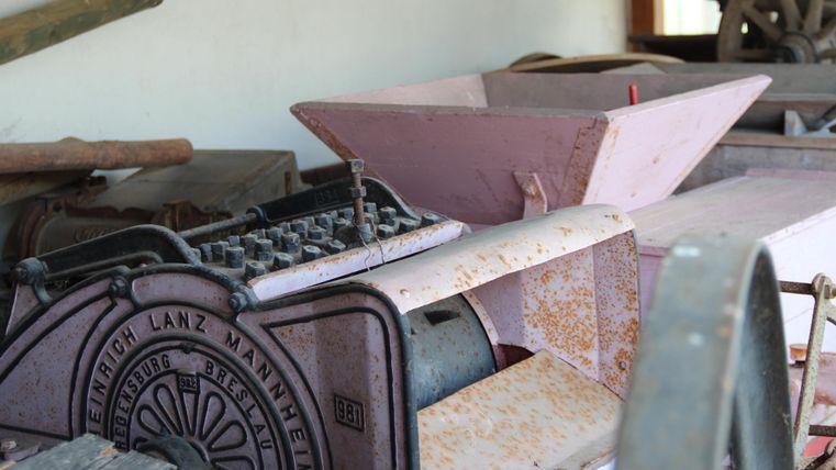 An old agricultural machine made of metal, rusty and worn out. In the background, other farming tools can be seen.