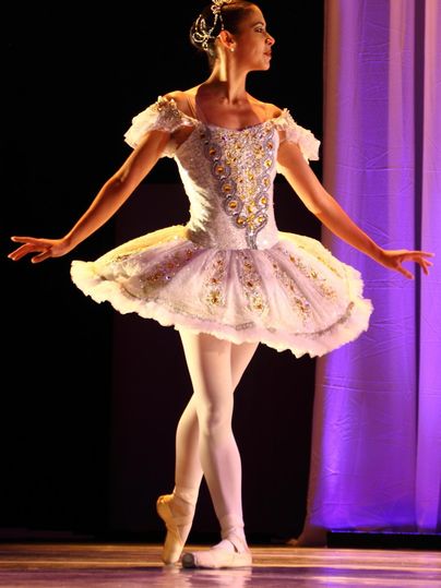 Une ballerine dans un costume élégant se tient sur scène. Son ensemble est blanc avec des ornements scintillants et l'éclairage crée une atmosphère festive.