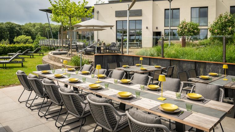 An elegant outdoor area with a long table set for a meal. In the background, a modern building and a well-kept garden can be seen.
