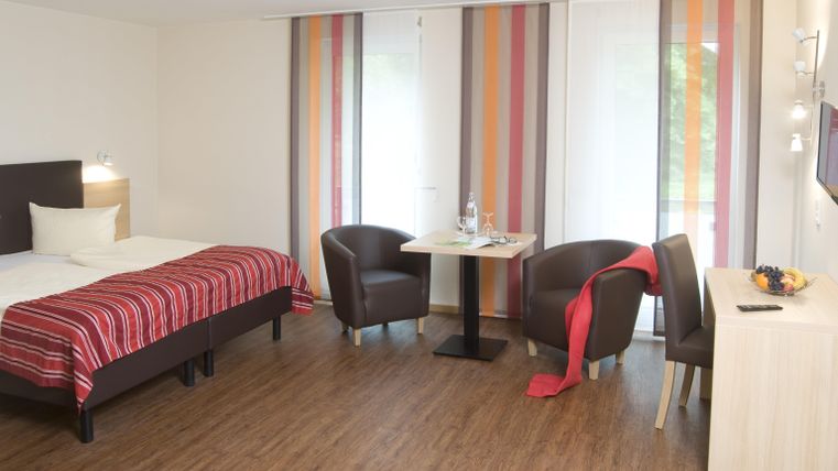 Ein modernes Zimmer mit einem Doppelbett und gemütlichen Sesseln. Große Fenster mit bunten Vorhängen sorgen für viel Licht.