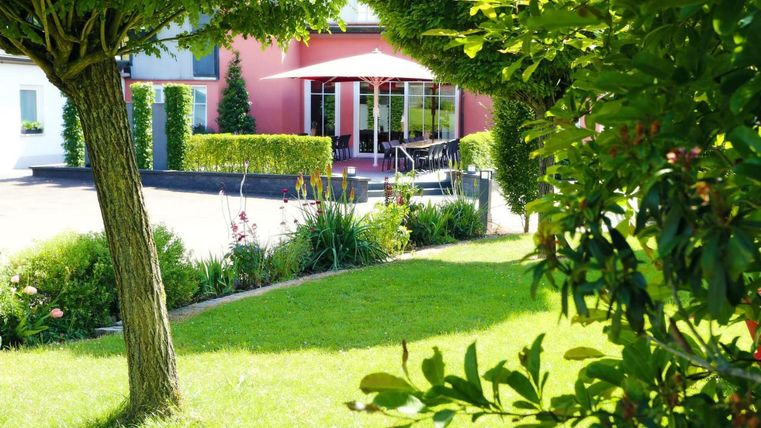 A beautiful garden with well-kept lawn and trees. In the background, there is a house with a terrace and a sun umbrella.