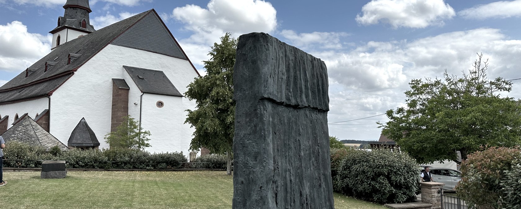 Kirche mit spitzem Turm, moderne Skulptur im Vordergrund, gr&uuml;ner Rasen, B&auml;ume und blauer Himmel mit Wolken.