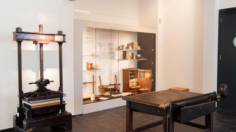 Une pièce avec une presse à imprimer sur le côté gauche et une table sur le côté droit. À l'arrière-plan, des vitrines avec des objets historiques sont visibles.