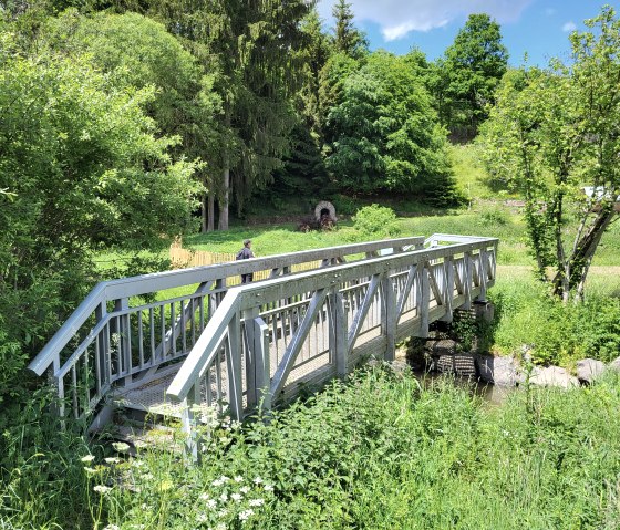 Een kleine houten brug leidt over een beek in een groen, bebost landschap. Op de achtergrond is een wandelaar te zien., © Tourist-Information Islek, Ingrid Wirtzfeld