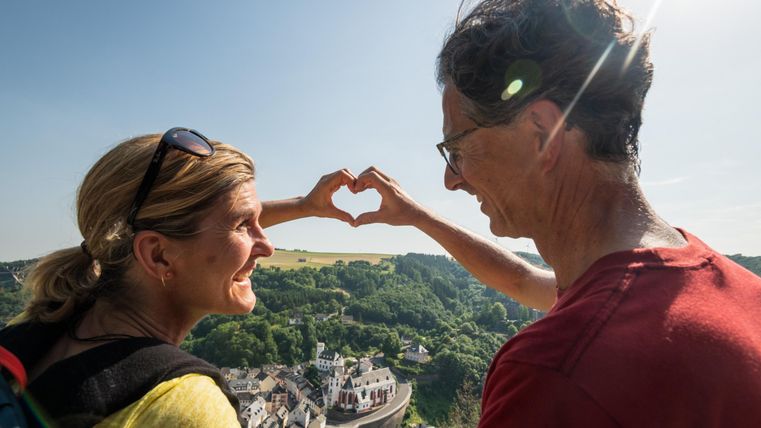A couple stands on a viewpoint and forms a heart with their hands. In the background, there is a picturesque landscape with a city and hills.