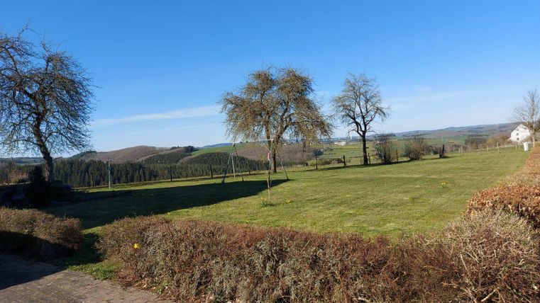 A wide landscape with lush green and a few trees. The sky is clear and blue, creating a sunny atmosphere.