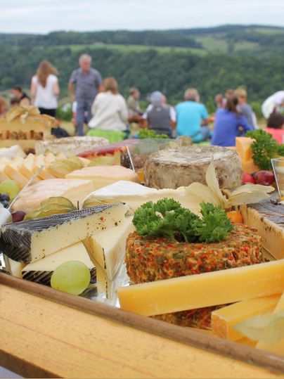 Een rijkelijk kaasplankje met verschillende kaassoorten en verse druiven. Op de achtergrond zijn veel mensen te zien in een pittoresk landschap.