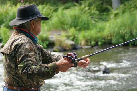 An older man is fishing at the riverbank. He is wearing a camouflage jacket and a hat while he focuses on fishing with the rod.