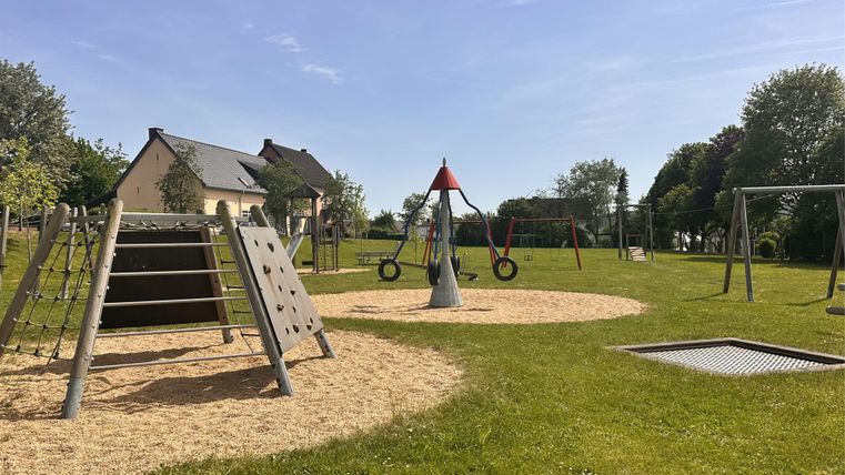 Ein Spielplatz mit verschiedenen Spielgeräten wie Rutschen und Schaukeln. Die Umgebung ist grün und der Himmel ist blau.