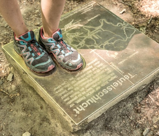 Une personne portant des chaussures de sport color&eacute;es se tient sur un panneau d'information dans la for&ecirc;t. Le panneau montre une carte et le texte 'Teufelsschlucht' (gorge du diable)., &copy; Felsenland S&uuml;deifel Tourismus GmbH