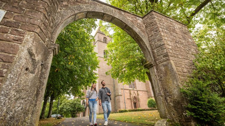 A romantic couple walks through an old stone gate. In the background, you can see a historic building environment with trees.