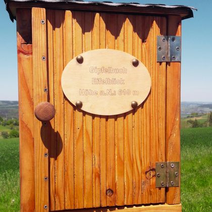 Eine Holzhütte mit einer ovalen Plakette. Der Hintergrund zeigt eine grüne Wiese und einen klaren Himmel.