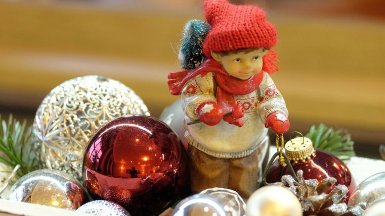 A small Christmas figure in a red hat stands among colorful ornaments and pine branches. The scene exudes a festive atmosphere.