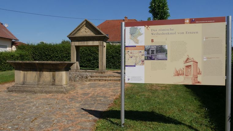 Ein historisches Denkmal mit einer Steinsäule und einem Informationsschild steht in einer grünen Umgebung. Im Hintergrund sind Häuser und ein blauer Himmel zu sehen.