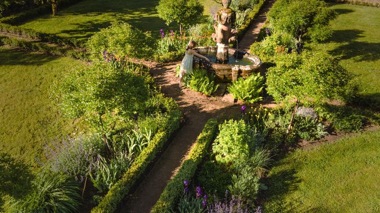 Ein schöner Garten mit einer Statue in der Mitte und vielen Pflanzen und Blumen. Der Garten ist von grünen Bäumen und Sträuchern umgeben.