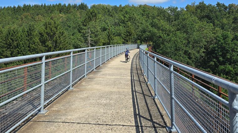 A wide bridge spans a green landscape. A cyclist is riding along the path toward the woods.