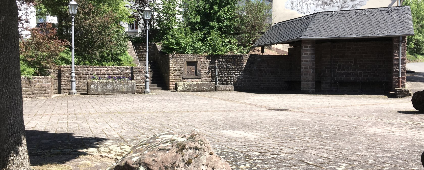 Place de village pav&eacute;e &agrave; Gransdorf avec des arbres, des lanternes et un b&acirc;timent avec une peinture murale. La place est entour&eacute;e de nature et de murs en pierre., &copy; TI Bitburger Land