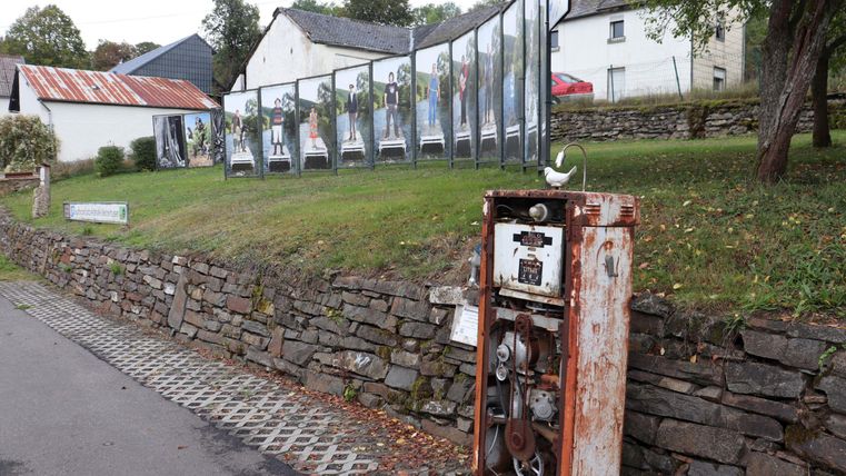 Eine alte, rostige Zapfsäule steht an einer Steinmauer. Im Hintergrund sind Fotos in großen Rahmen aufgestellt, die eine Ausstellung zeigen.