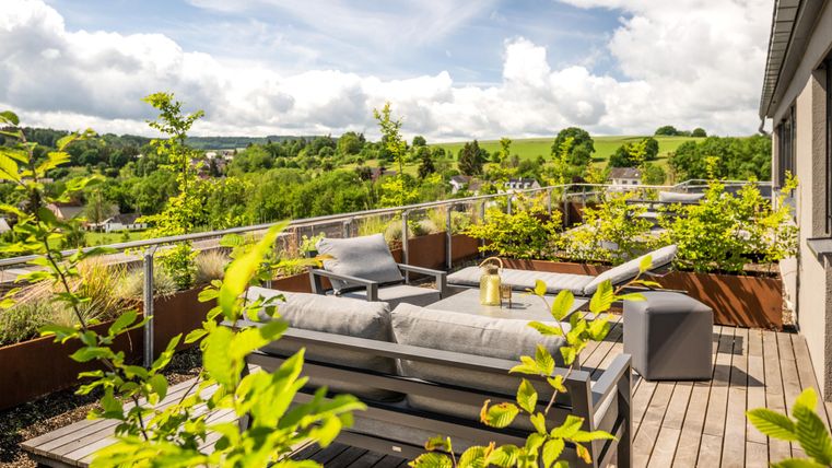 A modern rooftop terrace with comfortable seating and plenty of green plant growth. The view extends over an idyllic landscape under a bright sky.