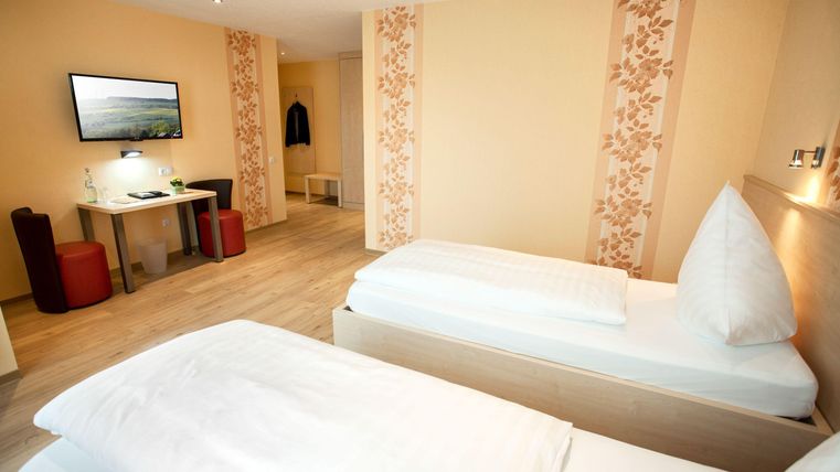 A bright room with two single beds and a desk. In the background, there is a television and cozy seating options.
