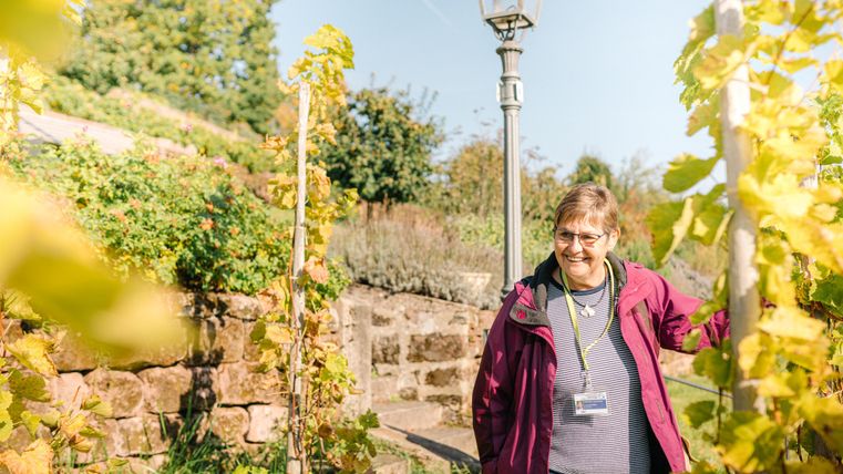 Une femme souriante se tient dans un jardin avec des vignes. En arrière-plan, on voit des plantes vertes et un lampadaire.