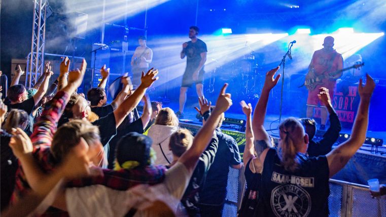 Een grote menigte juicht tijdens een live concert. Het podium is verlicht en de artiesten optreden met energie.