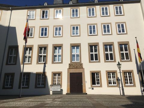 A historical building with several windows and two flags. The facade is bright and clear, surrounded by a square.