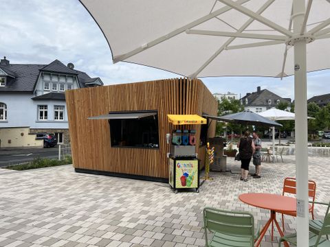 Een moderne houten stand met een drankuitgifte staat op een tegelpad. Op de achtergrond zijn parasols en enkele mensen te zien.