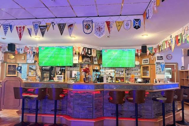 A sports bar with two large televisions broadcasting a soccer game. The walls are decorated with various flags, and there are accessible seating options.