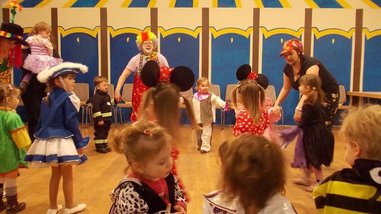 Une joyeuse mascarade pour enfants avec de nombreux enfants déguisés en costumes colorés. En arrière-plan, des clowns et des adultes participent, tandis que les enfants dansent et rient.
