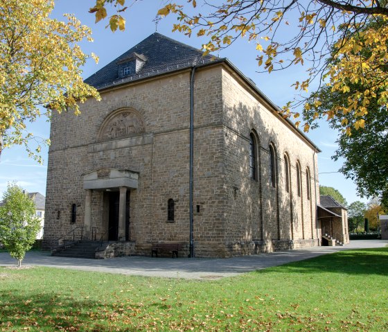 L'&eacute;glise Saint-Hubert de Wolsfeld, entour&eacute;e d'arbres automnaux, se trouve dans un parc verdoyant. Le b&acirc;timent est en pierre avec un toit &agrave; deux pentes., &copy; TI Bitburger Land