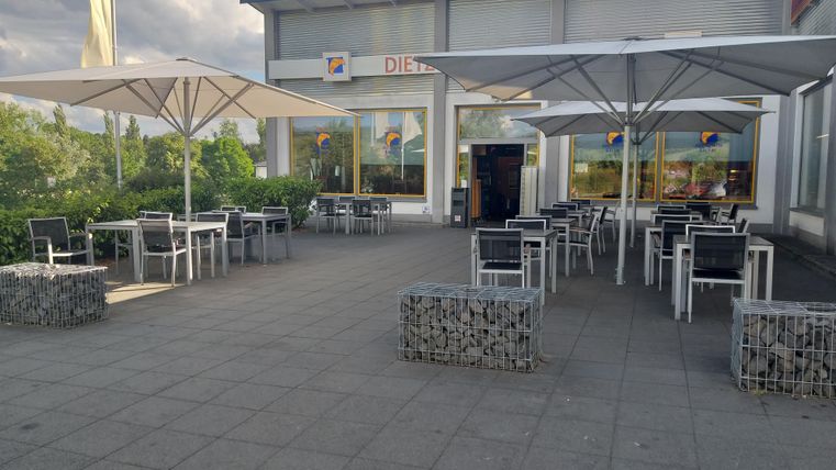 Une terrasse extérieure moderne avec des tables et des parasols. À l'arrière-plan se trouve un magasin avec de grandes fenêtres.