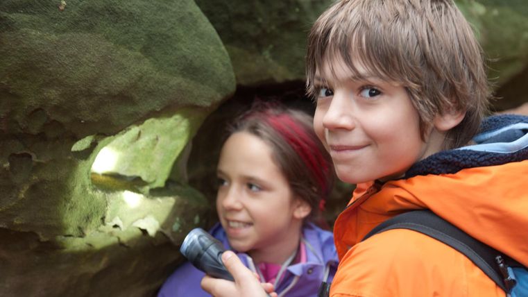 Twee kinderen verkennen een rotsformatie, terwijl een van hen de licht van een zaklamp vasthoudt. Ze lijken nieuwsgierig en enthousiast over hun avontuur.