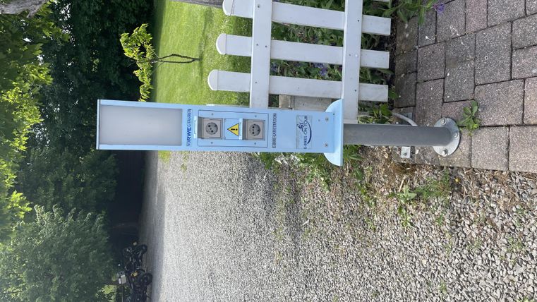 Een openbare laadstation voor elektrische voertuigen staat op een geplaveide weg. Op de achtergrond zijn bomen en een hek te zien.