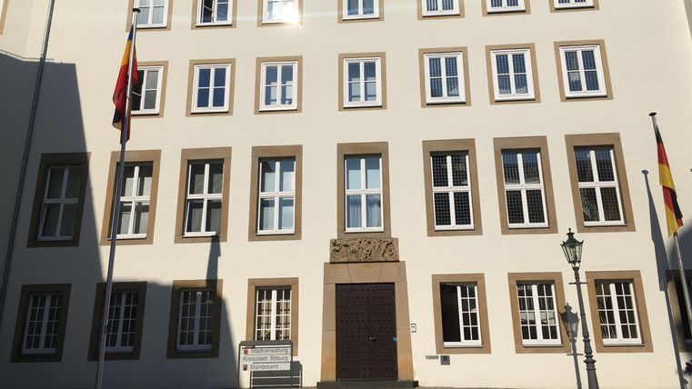 A historical building with several windows and two flags. The facade is bright and clear, surrounded by a square.